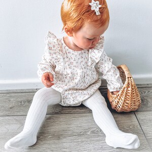 May include: A young child wearing a white floral print long-sleeved onesie with ruffles and white tights sits on a wood floor holding a wicker basket.