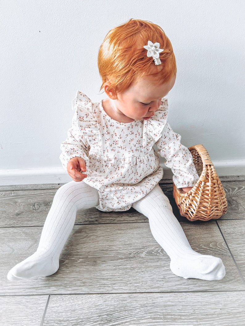May include: A young child wearing a white floral print long-sleeved onesie with ruffles and white tights sits on a wood floor holding a wicker basket.