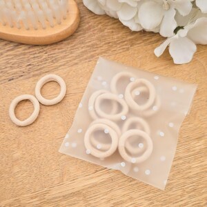 Puede incluir: Colecci&oacute;n de gomas para el pelo beige, algunas sueltas y otras en una bolsa de lunares, junto a un cepillo de pelo de madera y flores blancas. Las gomas para el pelo son de un material suave y el&aacute;stico.