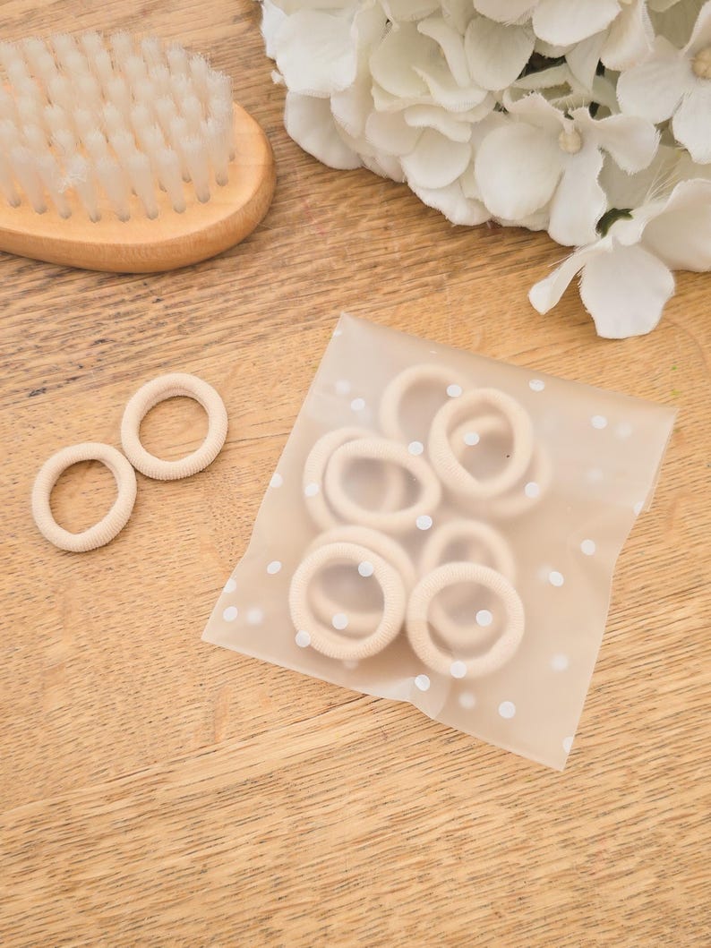 Puede incluir: Colecci&oacute;n de gomas para el pelo beige, algunas sueltas y otras en una bolsa de lunares, junto a un cepillo de pelo de madera y flores blancas. Las gomas para el pelo son de un material suave y el&aacute;stico.