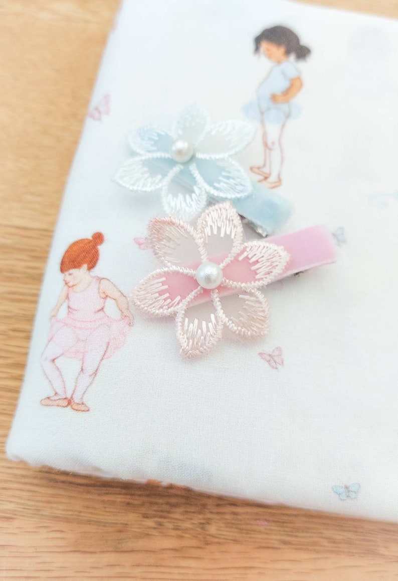 May include: Two hair clips with pink and blue velvet ribbon and a flower design. The flowers are made of sheer fabric with a pearl in the center.