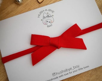 Puede incluir: Una tarjeta blanca con un lazo y cinta rojos, con el texto "Hush-a-bye Bay" y "Handmade with love for your little loves". La tarjeta está decorada con pequeños corazones de madera, campanas plateadas y copos de nieve dorados.