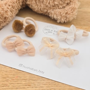 Puede incluir: Un conjunto de cinco gomas para el pelo sobre una tarjeta blanca con la etiqueta HUSH-A-BYE BAY. La colección incluye dos gomas con pompones marrones, dos gomas con lazos de cinta beige claro y una goma con un lazo blanco de tejido fino.