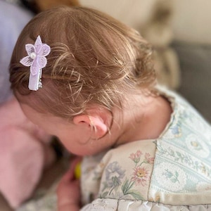May include: A young girl with blonde hair wearing a white dress with a floral pattern. She has a purple flower hair clip in her hair.