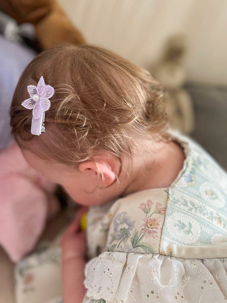 May include: A young girl with blonde hair wearing a white dress with a floral pattern. She has a purple flower hair clip in her hair.