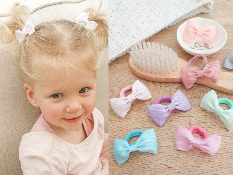May include: A collection of hair ties featuring small grosgrain ribbon bows in various pastel colours including pink, lavender, light blue, and mint green, each attached to a matching elastic band for securing hair in ponytails or pigtails.