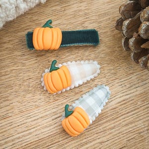 May include: Three hair clips with orange pumpkin accents. One clip has a dark green velvet base, while the other two have a gingham pattern in white and gray. The pumpkins have green stems. The clips are on a wooden surface.