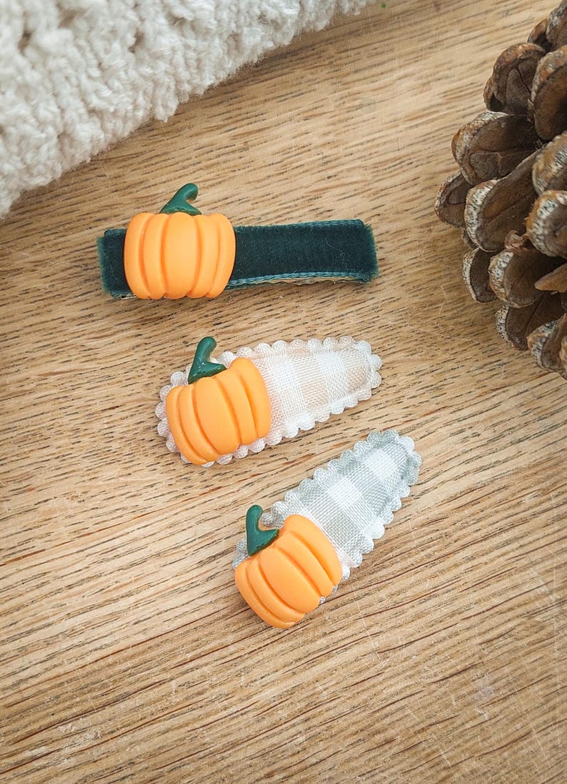 May include: Three hair clips with orange pumpkin accents. One clip has a dark green velvet base, while the other two have a gingham pattern in white and gray. The pumpkins have green stems. The clips are on a wooden surface.