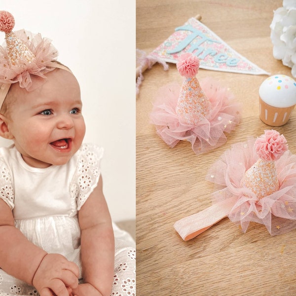 May include: A pink and white tulle headband with a glitter party hat. The headband is decorated with small white stars. The hat has a pink pom-pom on top.