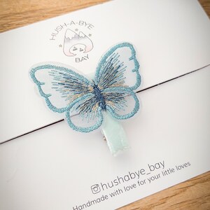 K&ouml;nnte beinhalten: Ein hellblauer Schmetterlings-Haarklammer mit wei&szlig;em Hintergrund. Die Klammer hat eine wei&szlig;e Samtr&uuml;ckseite und der Text "Hush-A-Bye Bay" ist auf den Hintergrund gedruckt. Der Text "Handmade with love for your little loves" ist ebenfalls auf den Hintergrund gedruckt.