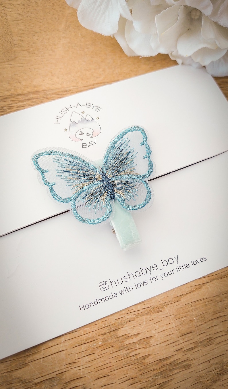 K&ouml;nnte beinhalten: Ein hellblauer Schmetterlings-Haarklammer mit wei&szlig;em Hintergrund. Die Klammer hat eine wei&szlig;e Samtr&uuml;ckseite und der Text "Hush-A-Bye Bay" ist auf den Hintergrund gedruckt. Der Text "Handmade with love for your little loves" ist ebenfalls auf den Hintergrund gedruckt.