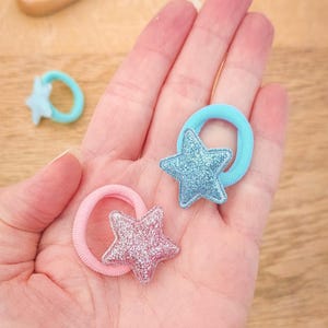 Pu&ograve; includere: Due elastici per capelli con ciondoli a forma di stella glitterata. Un elastico &egrave; azzurro chiaro e l'altro &egrave; rosa.