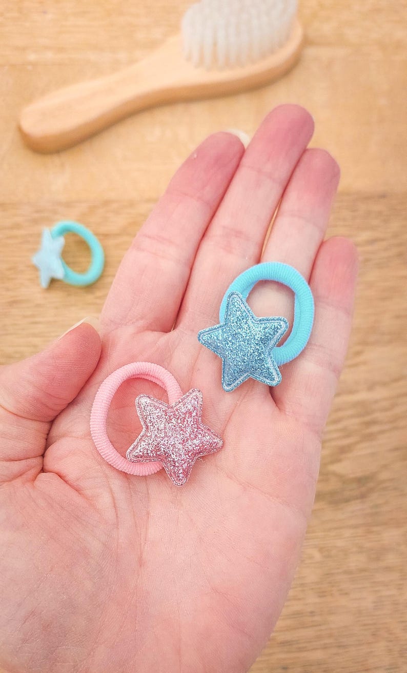 Pu&ograve; includere: Due elastici per capelli con ciondoli a forma di stella glitterata. Un elastico &egrave; azzurro chiaro e l'altro &egrave; rosa.
