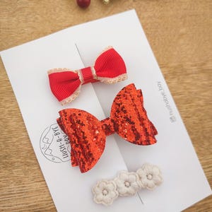 May include: Three red hair bows, one with glitter, one with a white trim, and one with a white and red trim. The bows are on a white card with the text "Hush-A-Bye  DIY" and "@hush_a_bye_toy".