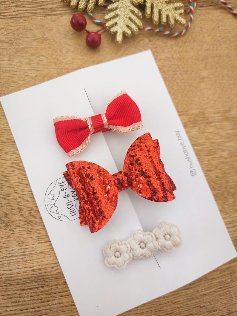 May include: Three red hair bows, one with glitter, one with a white trim, and one with a white and red trim. The bows are on a white card with the text "Hush-A-Bye  DIY" and "@hush_a_bye_toy".