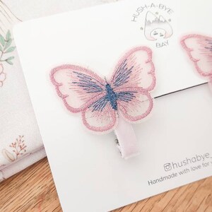 K&ouml;nnte beinhalten: Zwei rosa Schmetterlings-Haarklammern mit blauen Akzenten. Die Klammern befinden sich auf einer wei&szlig;en Karte mit dem Text "Hush-A-Bye Bay" und "@hushabye Handmade with love for you".