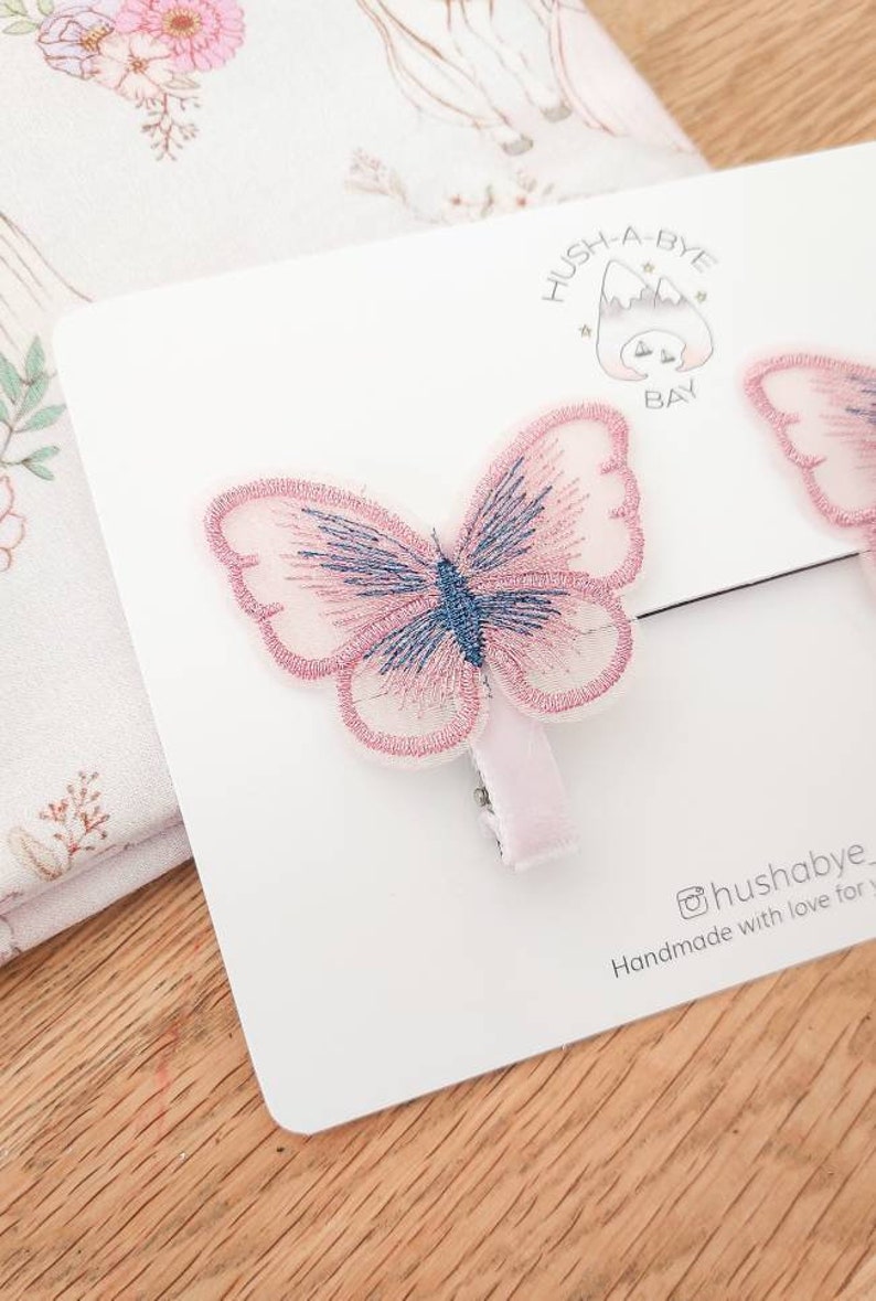 K&ouml;nnte beinhalten: Zwei rosa Schmetterlings-Haarklammern mit blauen Akzenten. Die Klammern befinden sich auf einer wei&szlig;en Karte mit dem Text "Hush-A-Bye Bay" und "@hushabye Handmade with love for you".