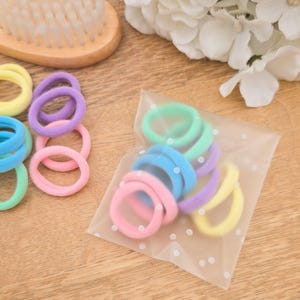 Puede incluir: Una colecci&oacute;n de gomas para el pelo de colores pastel en tonos rosa, azul, verde, morado y amarillo. Algunas est&aacute;n sueltas, otras en una bolsa transparente con lunares blancos. Un cepillo de madera y flores blancas en el fondo.