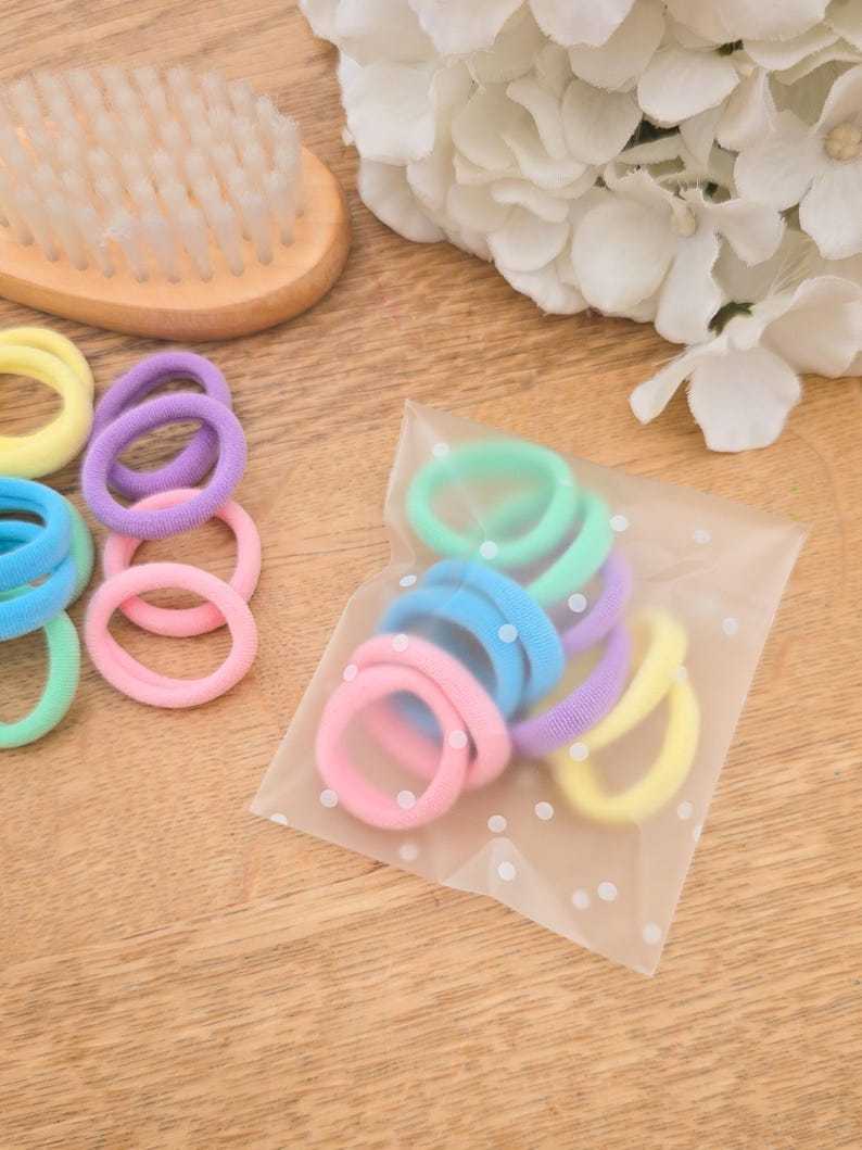 Puede incluir: Una colecci&oacute;n de gomas para el pelo de colores pastel en tonos rosa, azul, verde, morado y amarillo. Algunas est&aacute;n sueltas, otras en una bolsa transparente con lunares blancos. Un cepillo de madera y flores blancas en el fondo.