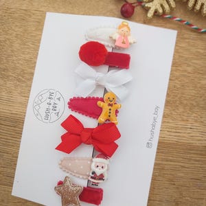 May include: Set of 7 Christmas-themed hair clips. The clips feature a variety of festive designs, including a gingerbread man, Santa Claus, a star, a bow, and an angel. The clips are all red and white.