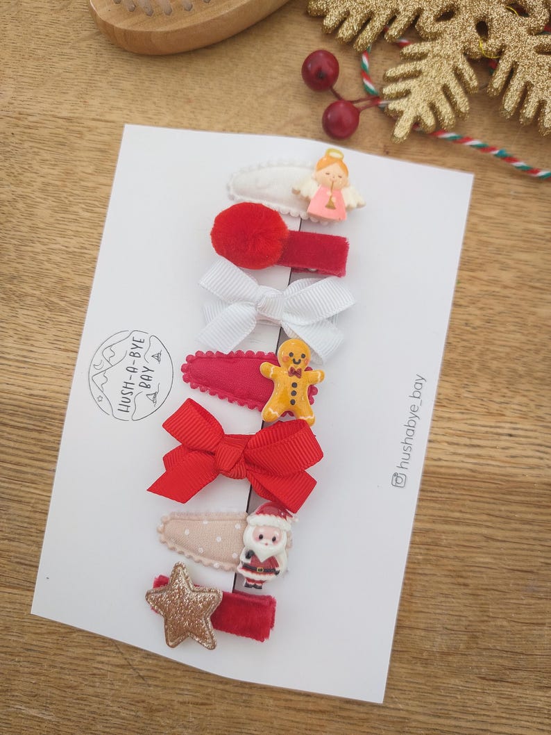 May include: Set of 7 Christmas-themed hair clips. The clips feature a variety of festive designs, including a gingerbread man, Santa Claus, a star, a bow, and an angel. The clips are all red and white.
