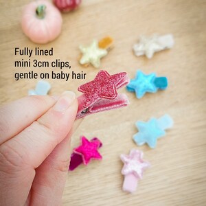 May include: A set of 10 mini star-shaped hair clips, each measuring 3 cm. The clips are made of velvet and have a glittery finish. The colors include pink, blue, and gold.  Fully lined mini 3cm clips, gentle on baby hair.