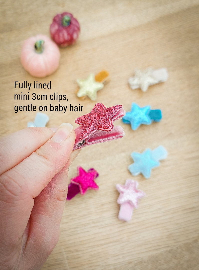 May include: A set of 10 mini star-shaped hair clips, each measuring 3 cm. The clips are made of velvet and have a glittery finish. The colors include pink, blue, and gold.  Fully lined mini 3cm clips, gentle on baby hair.