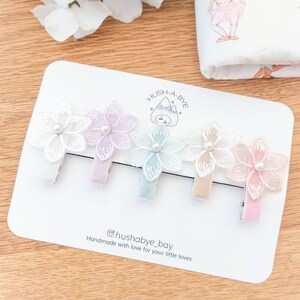 May include: Set of four pastel colored hair clips with delicate lace flowers and pearl accents. The clips are arranged on a white card with the text "Hush-A-Bye" and "@hushabye_bay Handmade with love for your little loves".