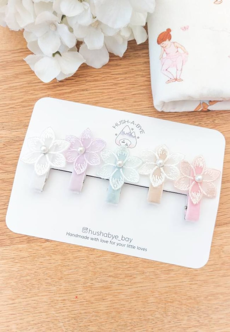 May include: Set of four pastel colored hair clips with delicate lace flowers and pearl accents. The clips are arranged on a white card with the text "Hush-A-Bye" and "@hushabye_bay Handmade with love for your little loves".