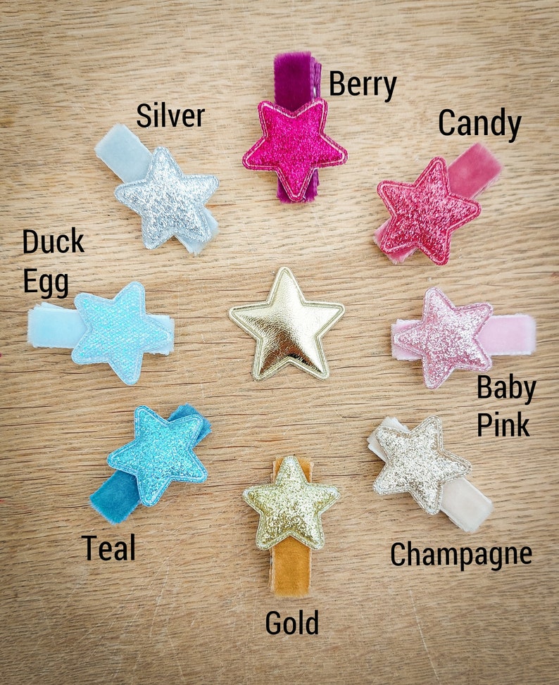 May include: Ten star-shaped hair clips in various colors and textures, including silver, teal, gold, berry, candy, baby pink, duck egg, and champagne. The clips are arranged on a wooden surface.