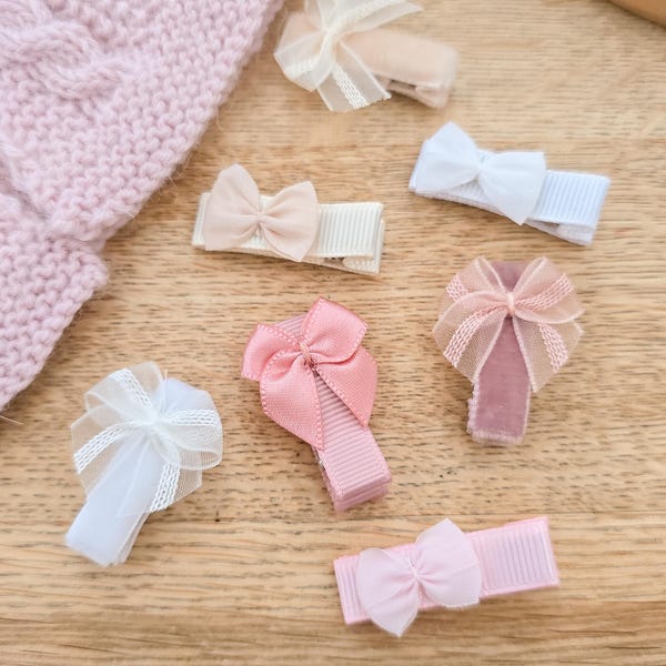 May include: A collection of seven hair clips featuring small bows in various shades of pink, white, and beige. The clips are made with grosgrain ribbon, velvet, and sheer mesh fabric, each topped with a delicate bow detail in matching colours.