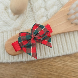 Puede incluir: Un lazo para el pelo a cuadros rojos, verdes y dorados. El lazo est&aacute; sujeto a un cepillo de pelo de madera. Una bellota de madera con una tapa dorada con purpurina est&aacute; en el fondo. El fondo es una superficie blanca texturizada.