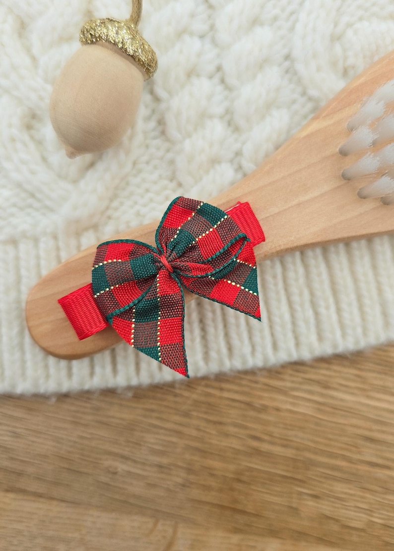 Puede incluir: Un lazo para el pelo a cuadros rojos, verdes y dorados. El lazo est&aacute; sujeto a un cepillo de pelo de madera. Una bellota de madera con una tapa dorada con purpurina est&aacute; en el fondo. El fondo es una superficie blanca texturizada.