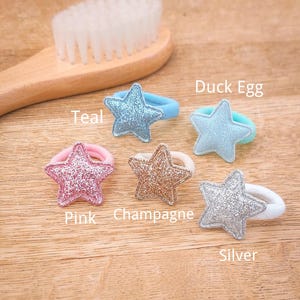Pu&ograve; includere: Cinque elastici per capelli a forma di stella in diversi colori: verde acqua, blu uovo di anatra, rosa, champagne e argento. Gli elastici per capelli sono su una superficie di legno.