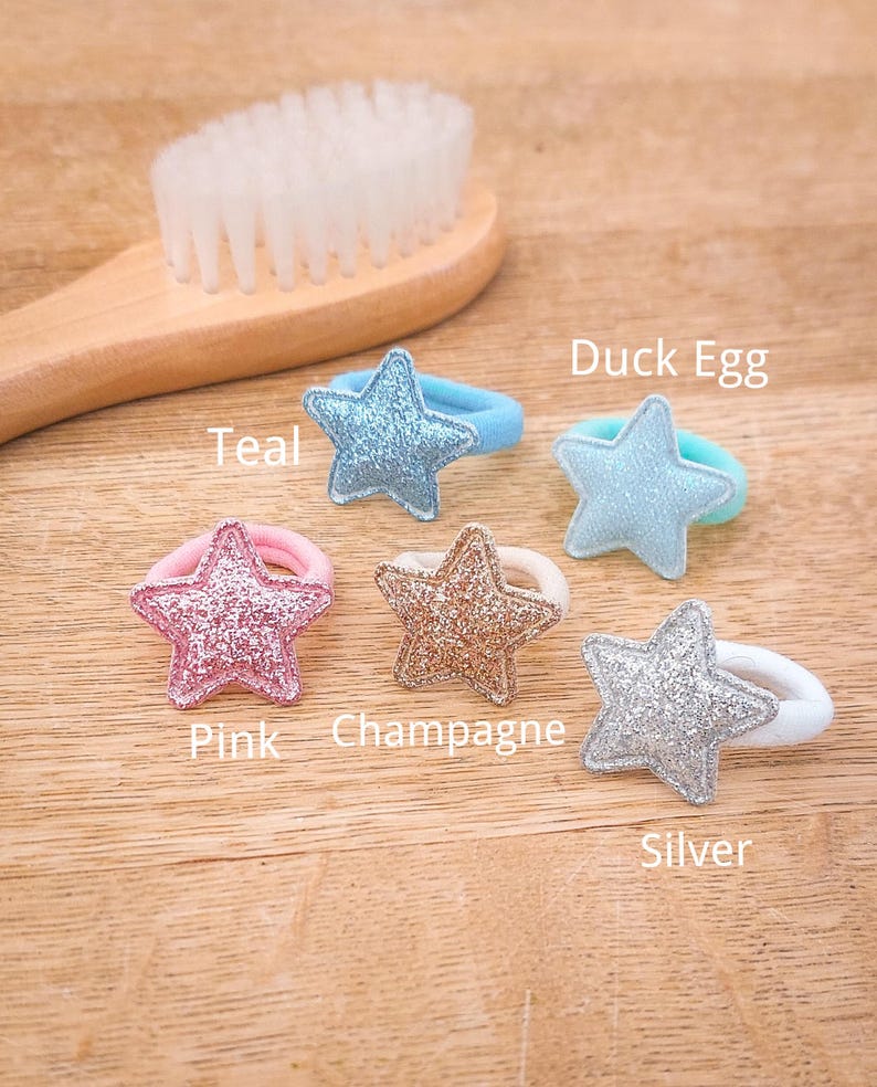 Pu&ograve; includere: Cinque elastici per capelli a forma di stella in diversi colori: verde acqua, blu uovo di anatra, rosa, champagne e argento. Gli elastici per capelli sono su una superficie di legno.