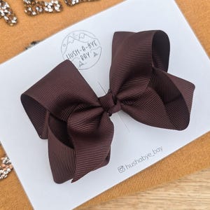 May include: A dark brown grosgrain ribbon hair bow attached to a white display card. The card features the logo HUSH-A-BYE BAY and the text hushabye_bay. The bow is centred on the card, which is placed on a mustard yellow fabric background.