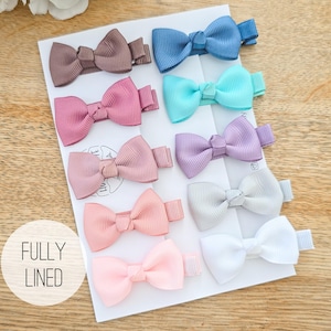 Peut inclure: Un ensemble de dix pinces à cheveux avec de petits nœuds en ruban gros-grain de différentes couleurs, dont marron, bleu, turquoise, rose, lavande, pêche, gris et blanc, sur un support blanc avec le texte FULLY LINED imprimé devant.