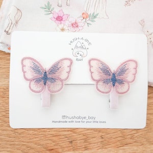K&ouml;nnte beinhalten: Zwei rosa Schmetterlingshaarspangen mit blauen Akzenten. Die Spangen befinden sich auf einer wei&szlig;en Karte mit dem Text "Hush-A-Bye Bay" und "@hushabye_bay Handmade with love for your little loves".