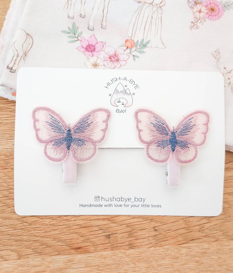 K&ouml;nnte beinhalten: Zwei rosa Schmetterlingshaarspangen mit blauen Akzenten. Die Spangen befinden sich auf einer wei&szlig;en Karte mit dem Text "Hush-A-Bye Bay" und "@hushabye_bay Handmade with love for your little loves".