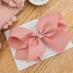 Puede incluir: Un lazo para el pelo de cinta de grosgrain color rosa empolvado sujeto a una tarjeta de presentación blanca con el texto HUSH-A-BYE BAY y hushabye_bay. El lazo tiene un centro anudado y bucles anchos y estructurados con textura acanalada.