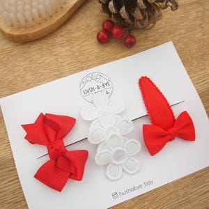May include: Three red and white hair clips. Two of the clips are red bows and one is a red alligator clip with a white lace flower. The clips are on a white card with the text "LUSH-A-BYE" and "@hushabye_bay".