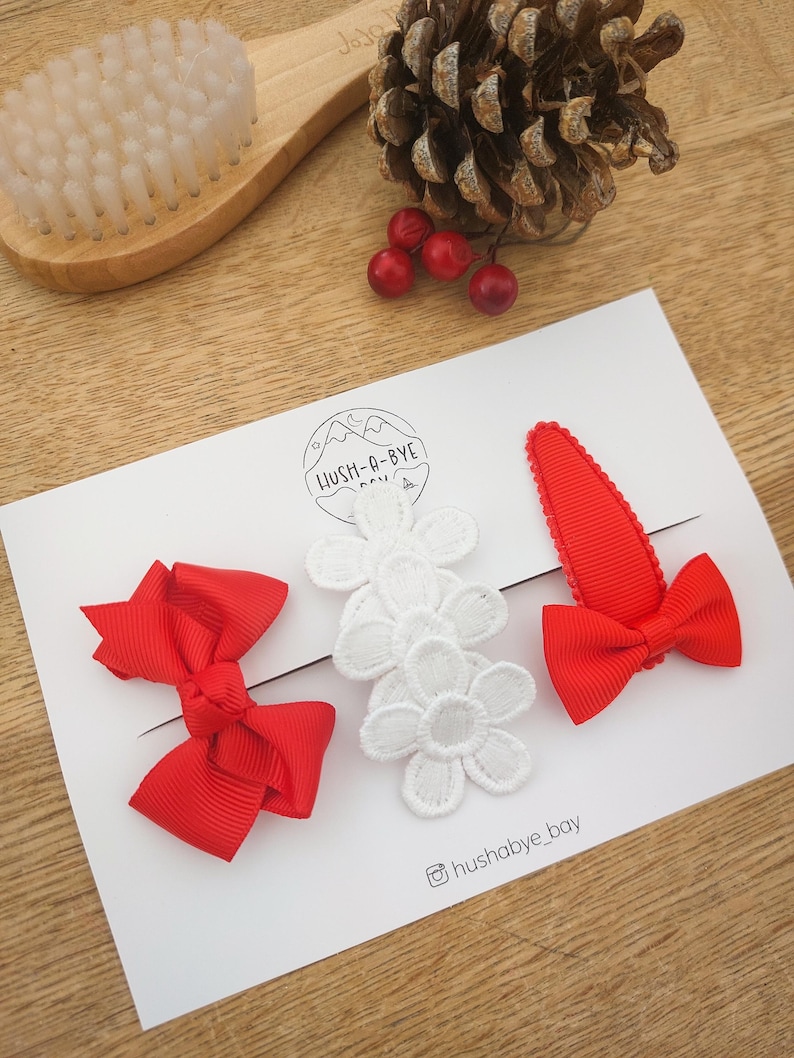 May include: Three red and white hair clips. Two of the clips are red bows and one is a red alligator clip with a white lace flower. The clips are on a white card with the text "LUSH-A-BYE" and "@hushabye_bay".