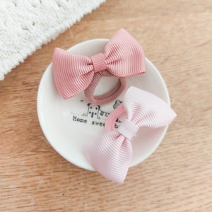 May include: Two hair ties with ribbed fabric bows in shades of dusty rose and pale pink. The bows are centred on elastic bands and are resting inside a small white ceramic dish featuring the printed text Home sweet home in black lettering.