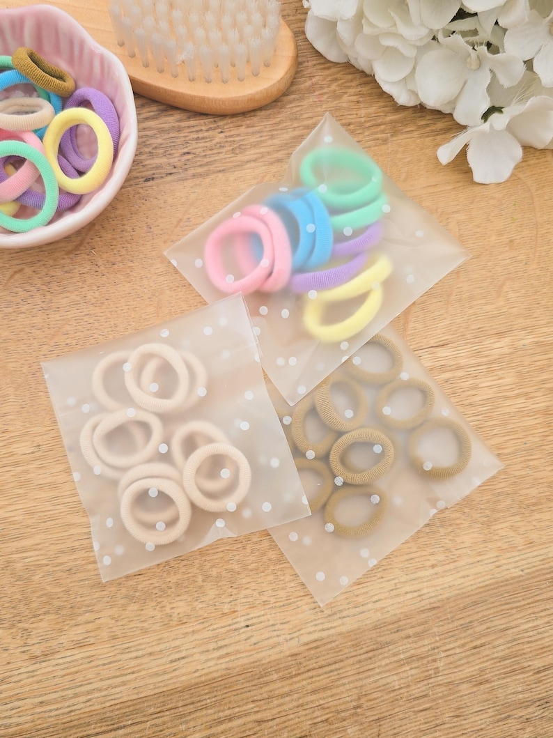 Puede incluir: Surtido de gomas para el pelo de colores pastel, incluyendo rosa, azul, verde, amarillo y morado. Algunas est&aacute;n en un cuenco peque&ntilde;o con forma de coraz&oacute;n, mientras que otras est&aacute;n empaquetadas en bolsas transparentes con lunares blancos. Un cepillo de madera y flores blancas en el fondo.