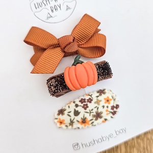 Könnte beinhalten: Ein Set aus drei Haarspangen auf einer weißen Präsentationskarte mit der Aufschrift HUSH-A-BYE BAY. Das Set enthält eine rostfarbene Schleife, eine dunkelbraune Glitzerspange mit einem orangefarbenen Kürbis und eine cremefarbene Spange mit Blumenmuster.