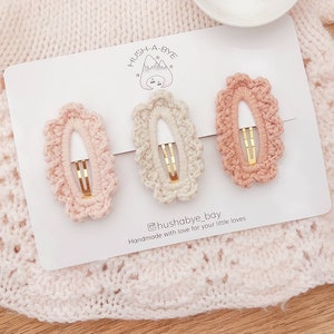 May include: Three crocheted hair clips in pink, beige, and peach colors. The clips are oval shaped with a gold metal clasp. The clips are displayed on a white card with the text "HUSH-A-BYE" and "@hushabye_bay Handmade with love for your little loves".