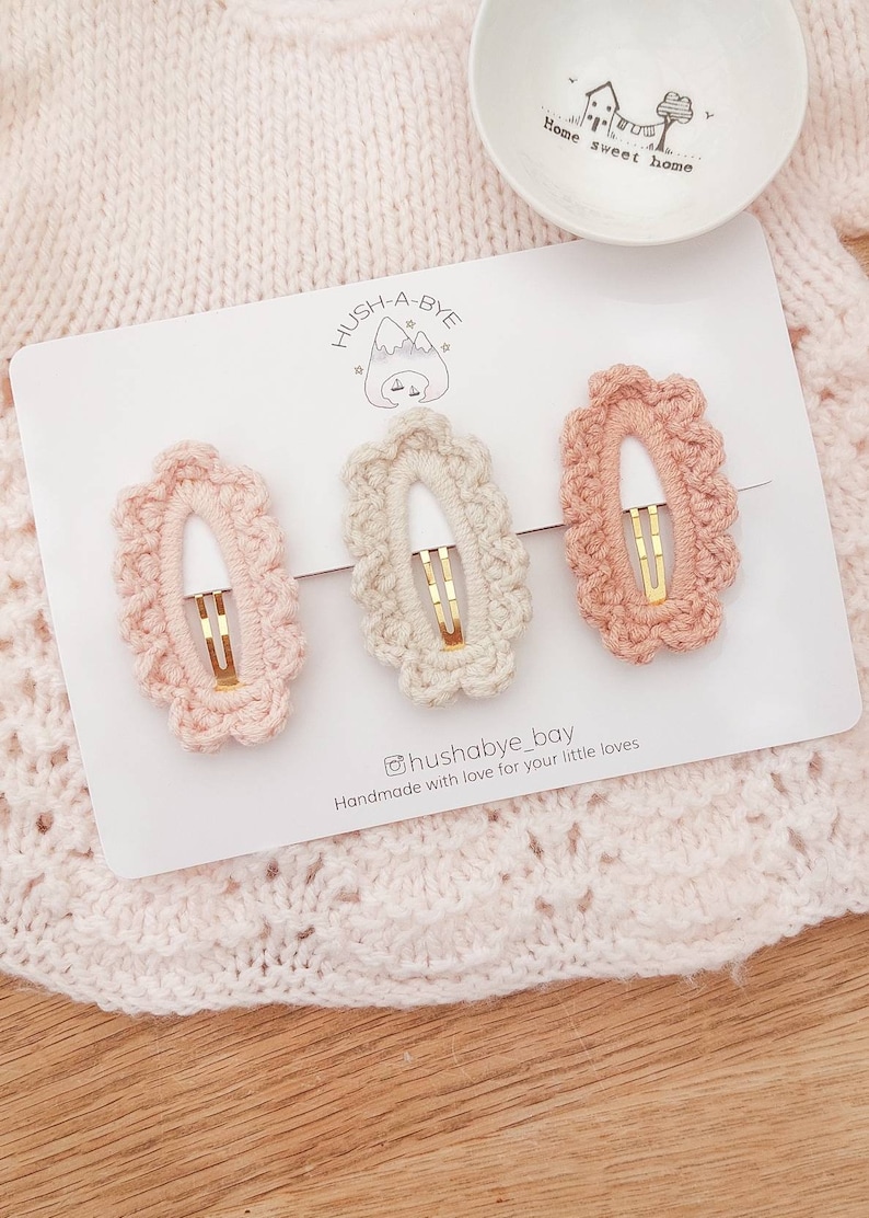 May include: Three crocheted hair clips in pink, beige, and peach colors. The clips are oval shaped with a gold metal clasp. The clips are displayed on a white card with the text "HUSH-A-BYE" and "@hushabye_bay Handmade with love for your little loves".