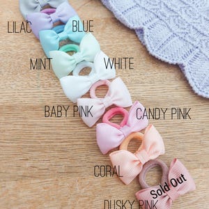 May include: A collection of hair ties with small grosgrain ribbon bows in various colours including grey, lilac, blue, mint, white, baby pink, candy pink, coral, and dusky pink. Each bow is attached to a matching elastic band for hair styling.