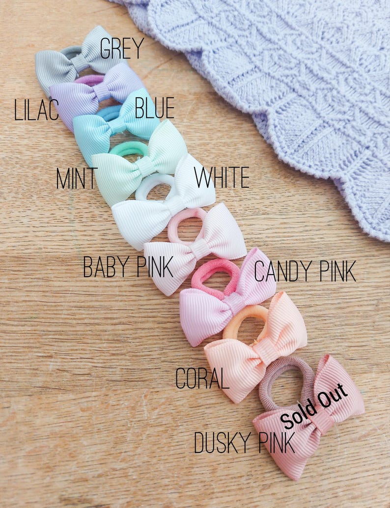 May include: A collection of hair ties with small grosgrain ribbon bows in various colours including grey, lilac, blue, mint, white, baby pink, candy pink, coral, and dusky pink. Each bow is attached to a matching elastic band for hair styling.