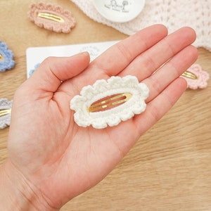 May include: A white crocheted hair clip with a gold metal bar. The clip is oval shaped with a scalloped edge.